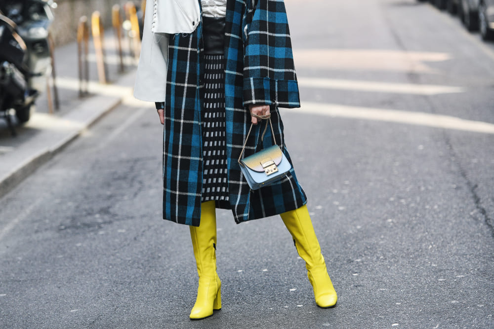 femeie imbracata cu palton in carouri albastre, fusta cu negru si alb si cizme inalte galbene